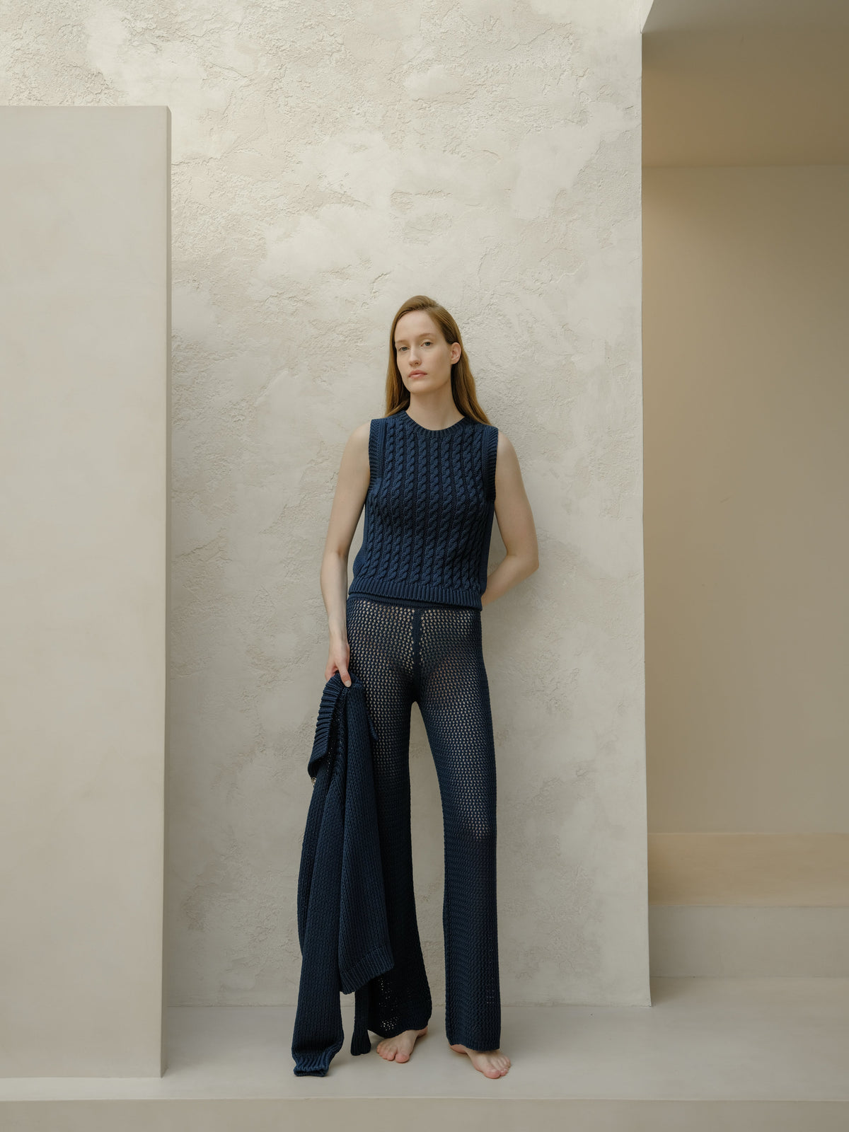Modella con blu navy knit sleeveless top e blu navy openwork pants holds a matching blu navy cardigan davanti a una parete chiara testurizzata