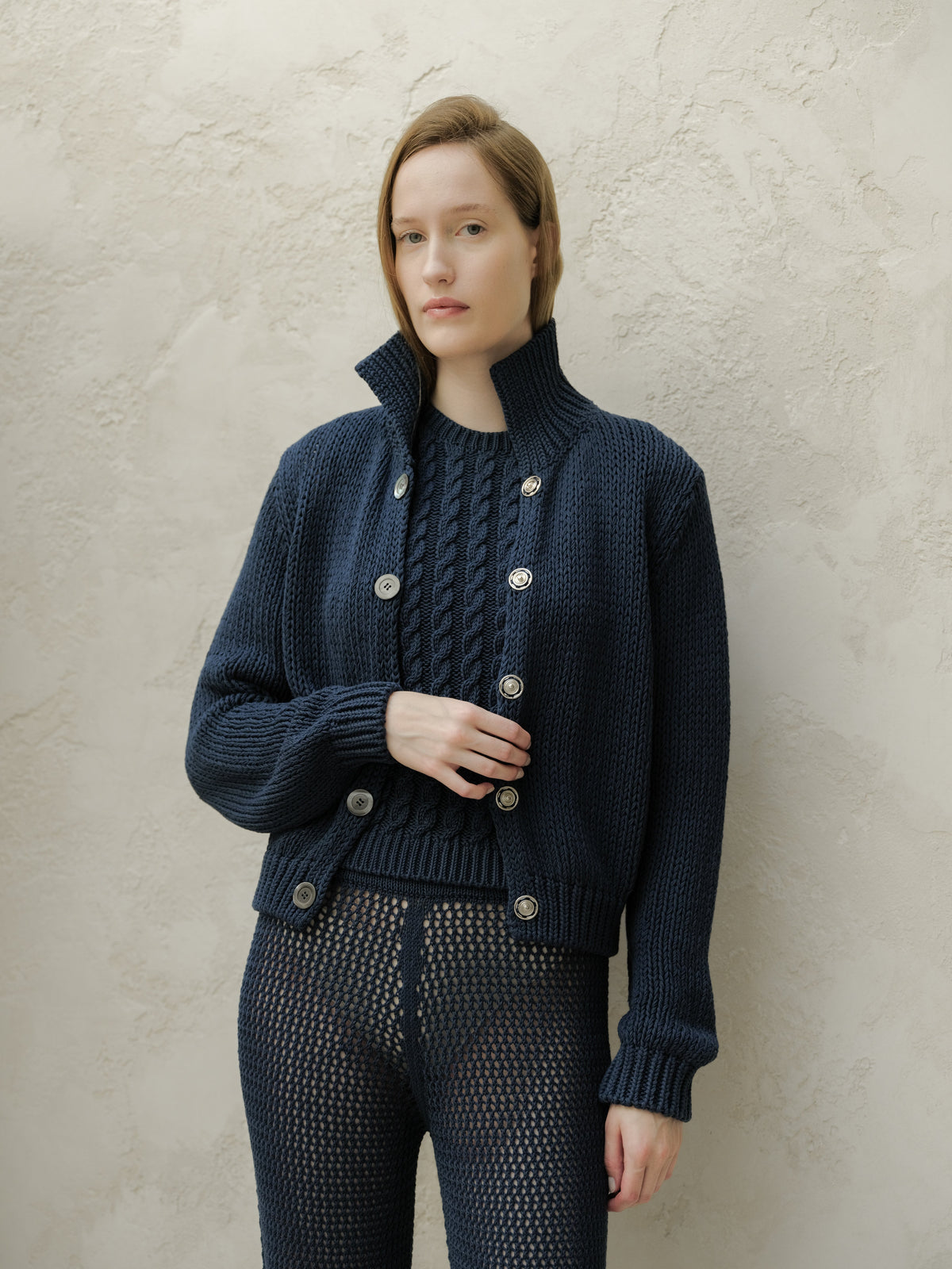 Donna con a blu navy chunky knit cardigan con a tall collar e large buttons stes against a texturosso beige wall.