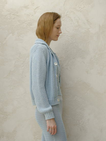 Side profile of a woman in a azzurro knitted bomber jacket con bianco buttons against a texturosso beige wall.