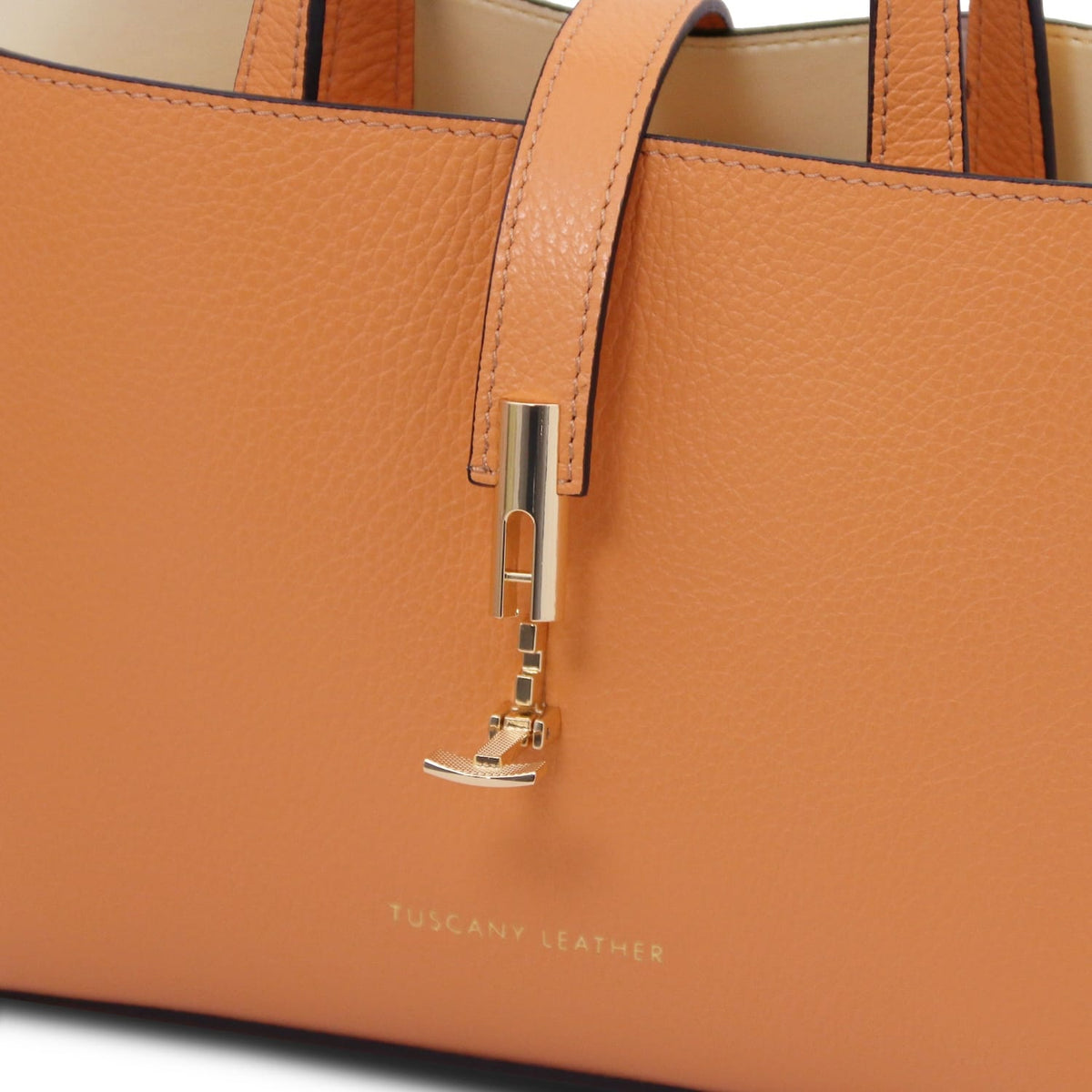 Close-up of tan pebbled leather tote with gold hardware and embossed "Tuscany Leather" branding on smooth leather surface.