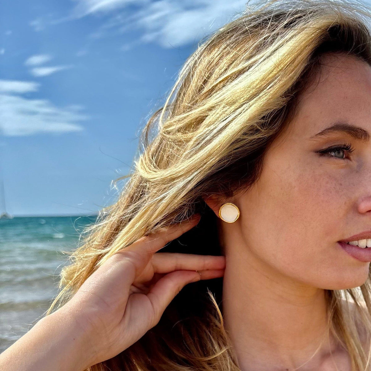 Camelia white ceramic stud earring with gold luster worn on a woman's ear at the beach