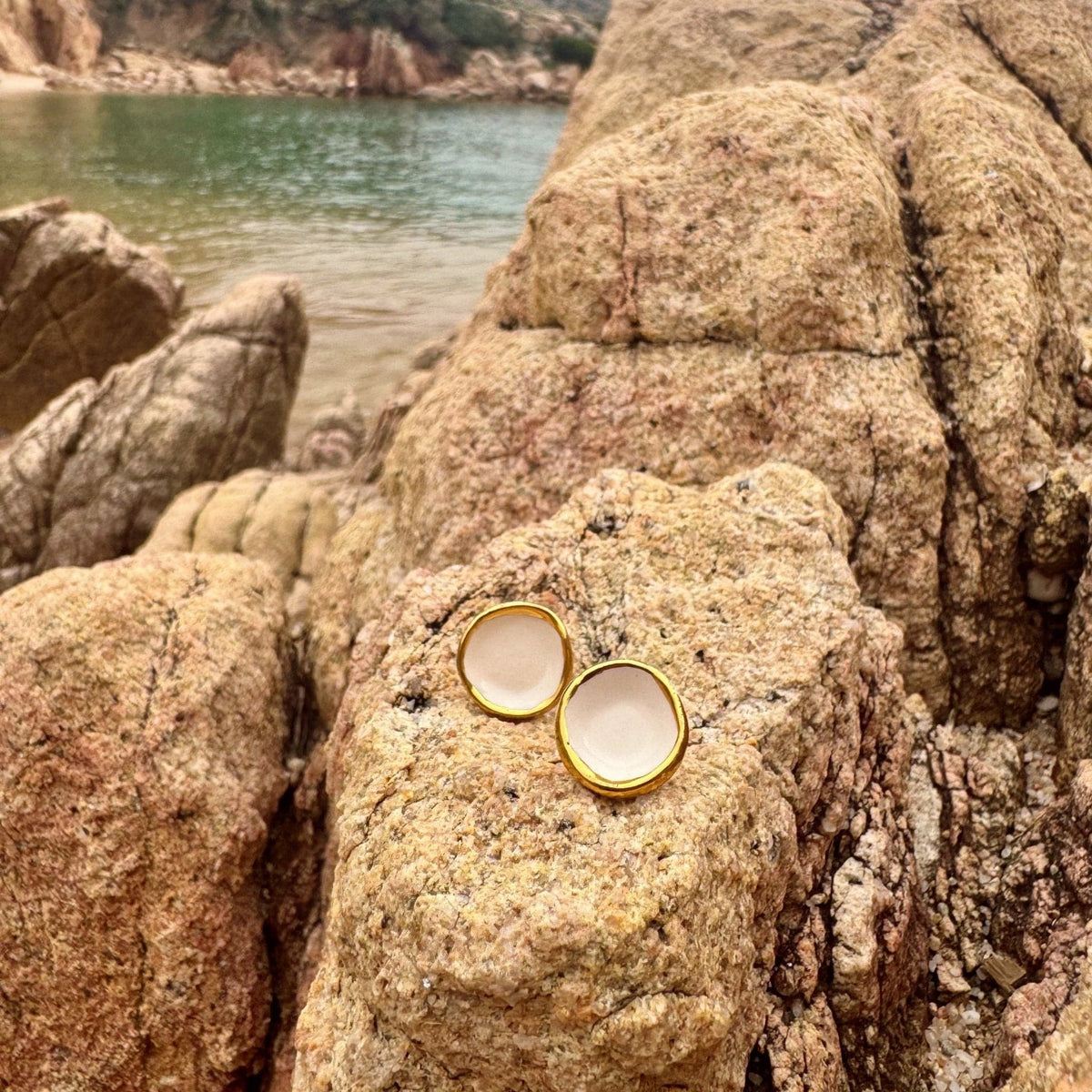 Camelia white ceramic stud earrings with gold trim resting on rugged coastal rocks