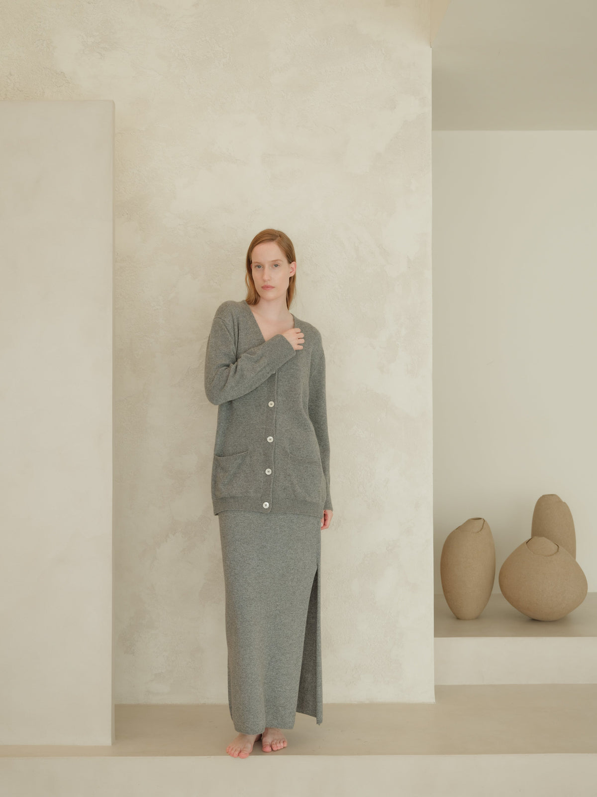 Front view of a woman in a gray knit cardigan with white buttons over a matching long skirt that features a front slit in a minimalist studio.