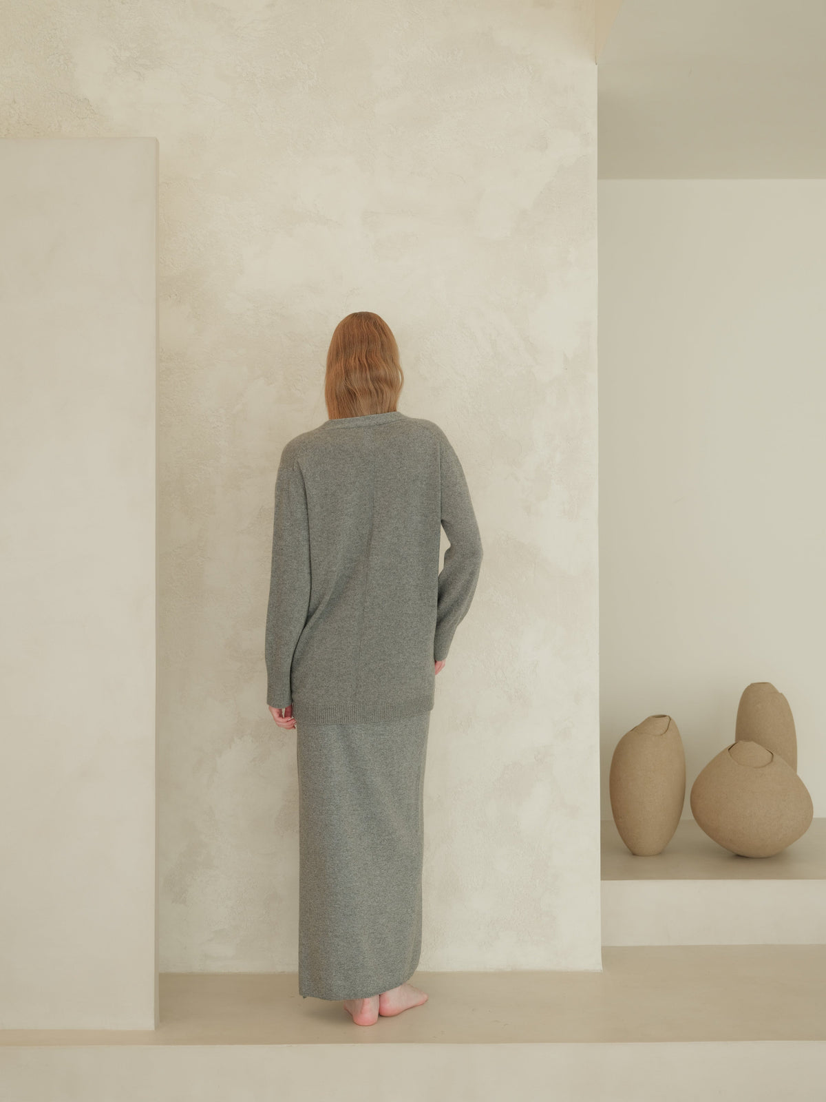 Back view of a model in a grey knit cardigan and long skirt standing against a textured wall