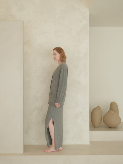 Side profile of a woman in a gray knit cardigan and matching long skirt, barefoot, against a textured beige backdrop with sculptural pots.
