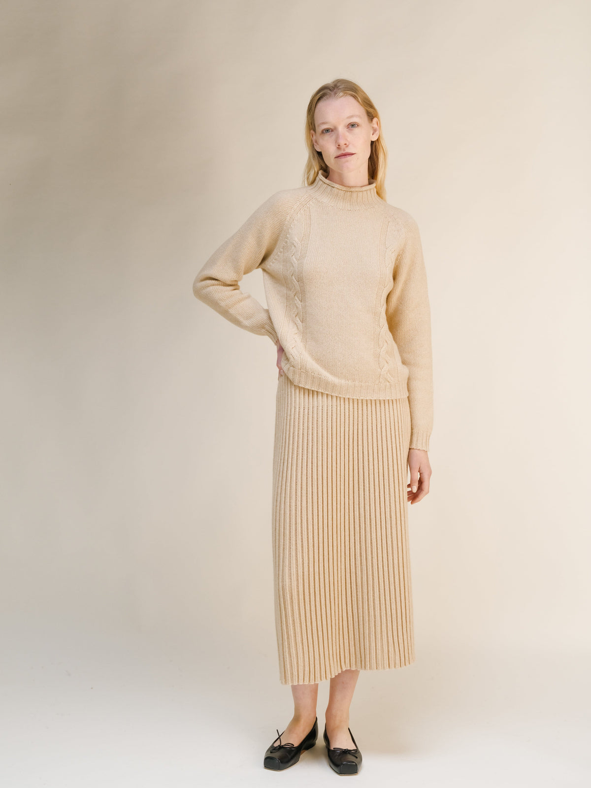 Model in a beige cable knit sweater and matching ribbed midi skirt against a neutral backdrop.