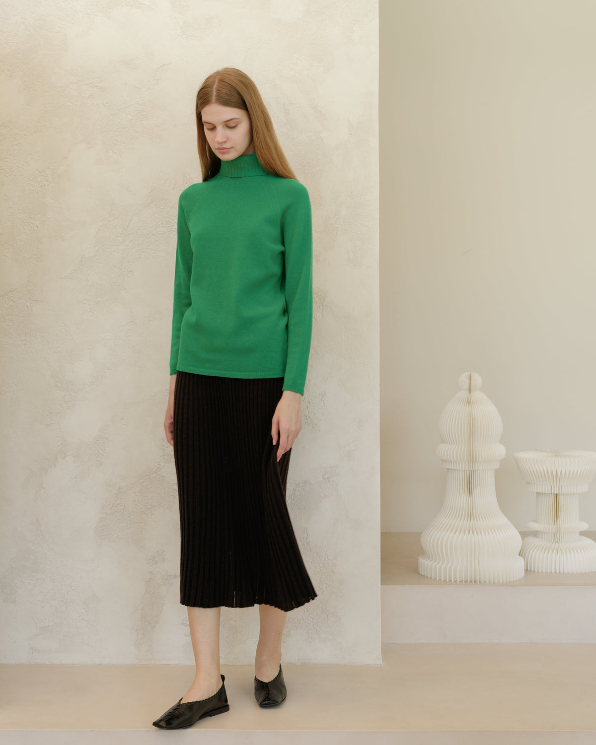 Model wearing bright green knit turtleneck and black midi pleated skirt against a textured beige wall.