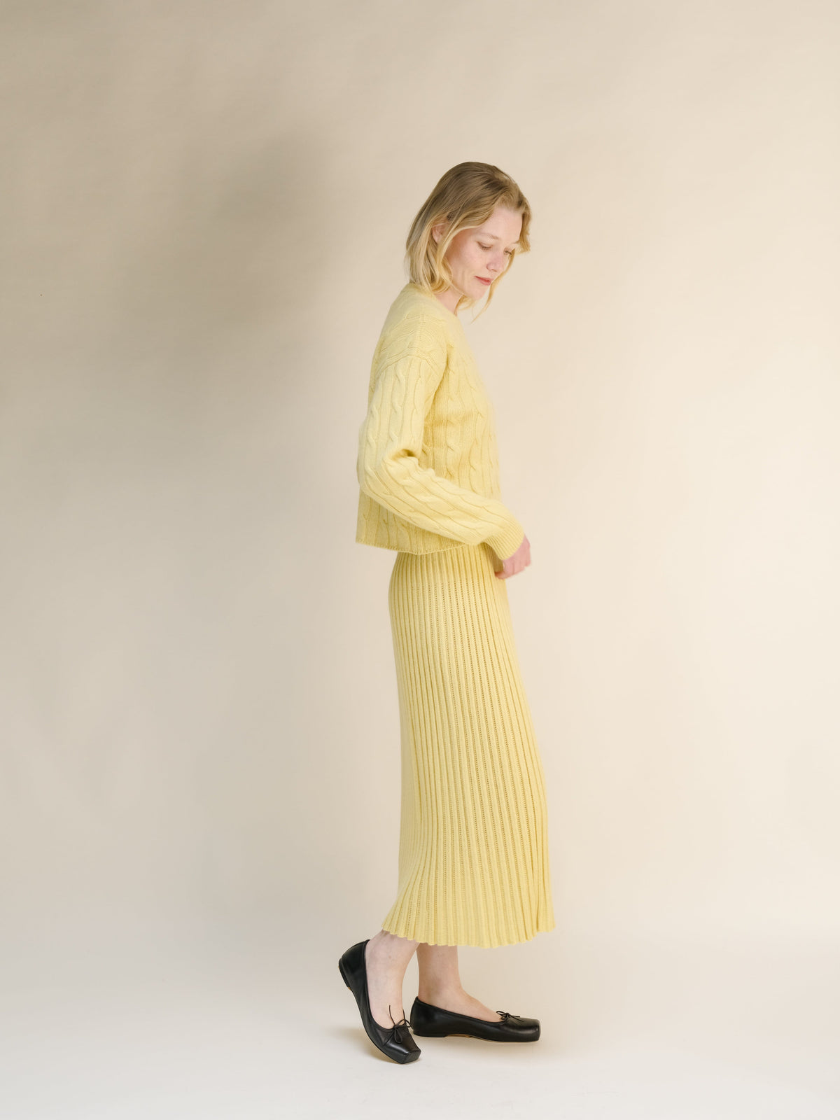 Side view of a woman wearing a pale yellow cashmere blend knit skirt with a matching sweater against a neutral backdrop.