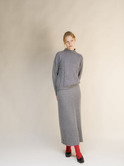 Full-length shot of a woman in a gray cable knit sweater and matching ribbed maxi skirt against a beige backdrop.