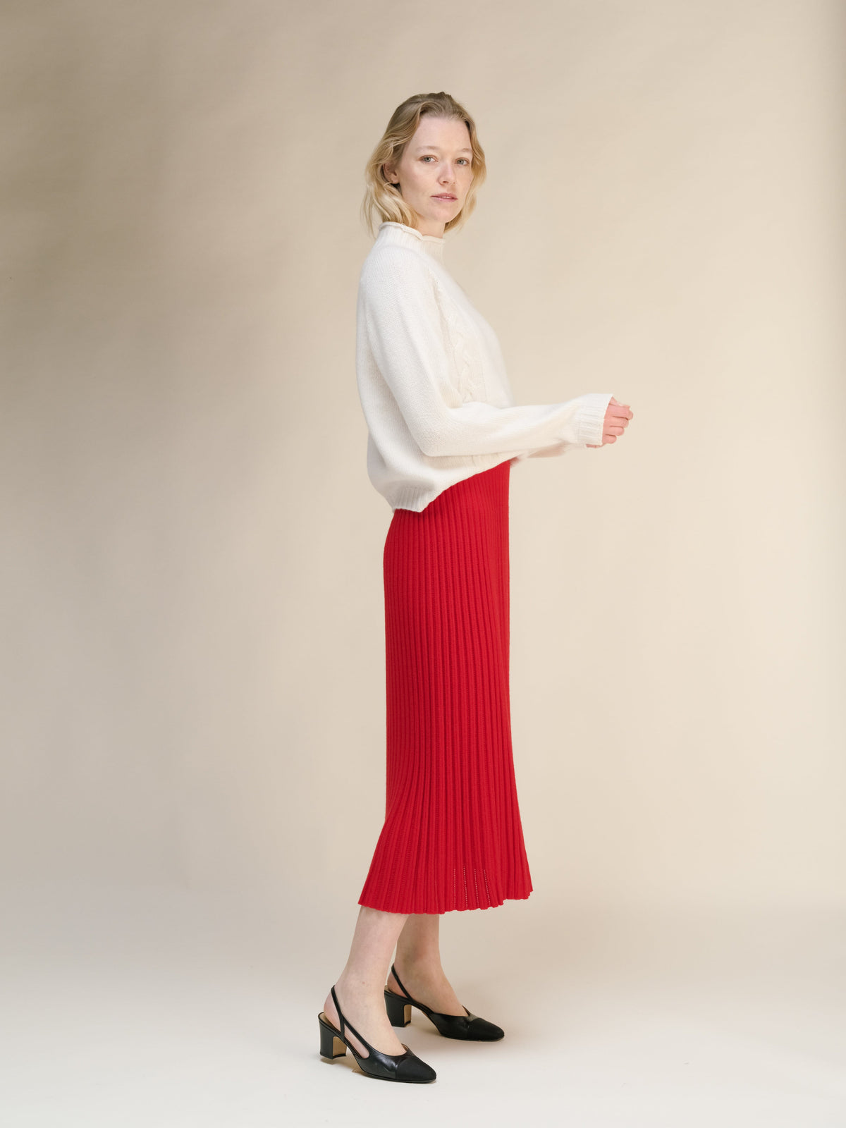 Side profile of a model wearing a cream knit sweater and red ribbed midi skirt with black heels.