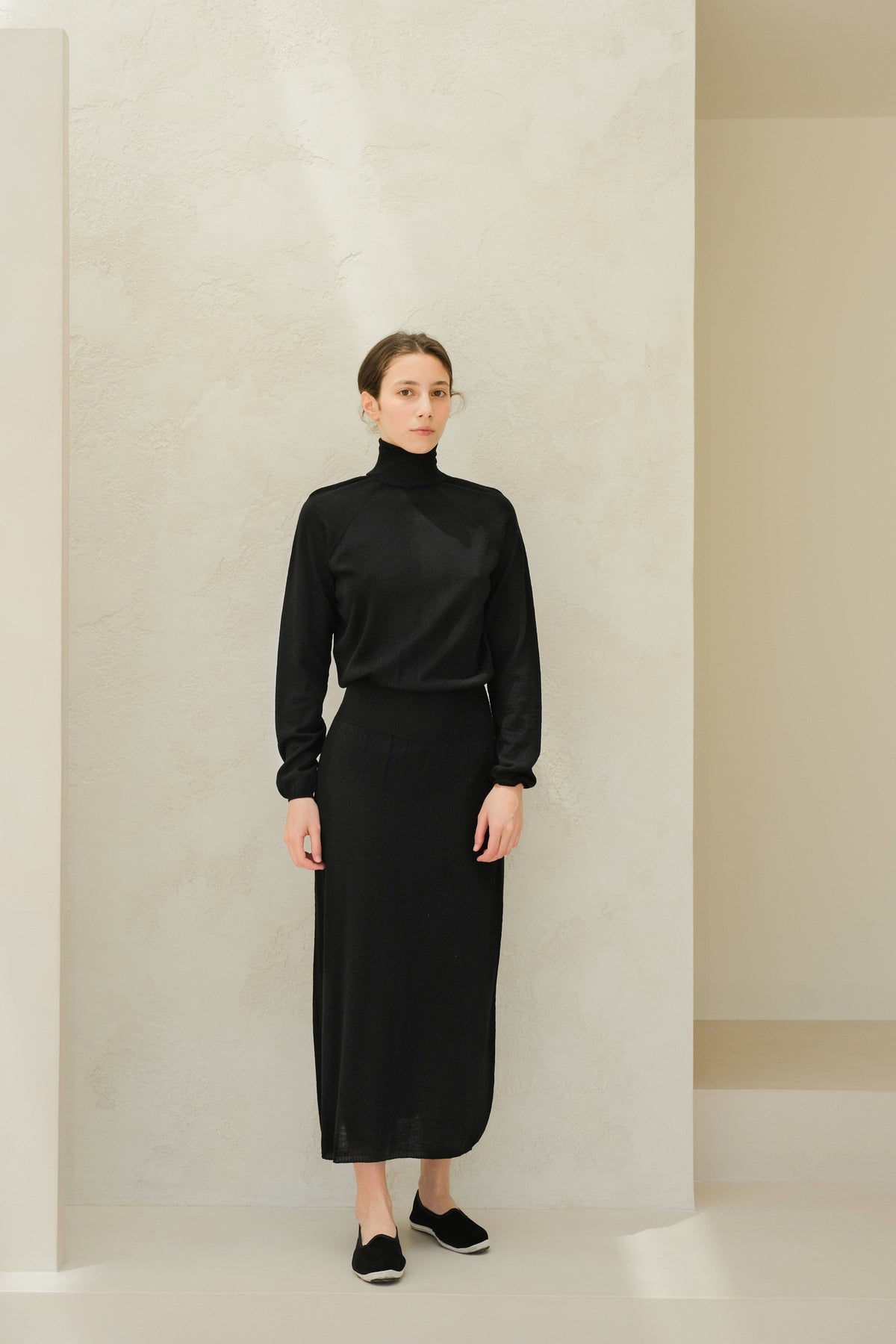 Model in a black 3D-knit merino wool dress with a turtleneck, full-length, standing against a textured wall.