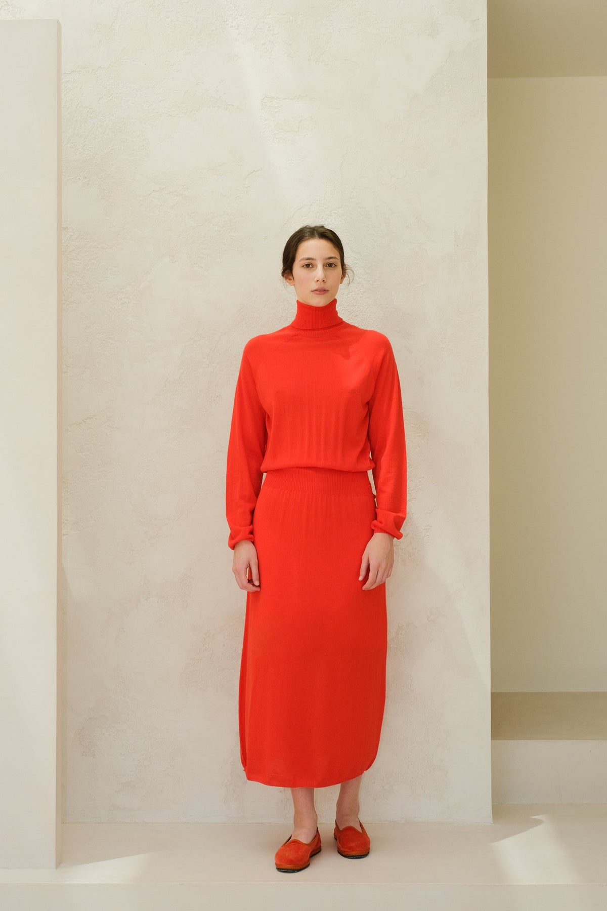 Model facing forward in a bright red 3D knit merino wool dress with a high neck and ribbed waist, midi-length.