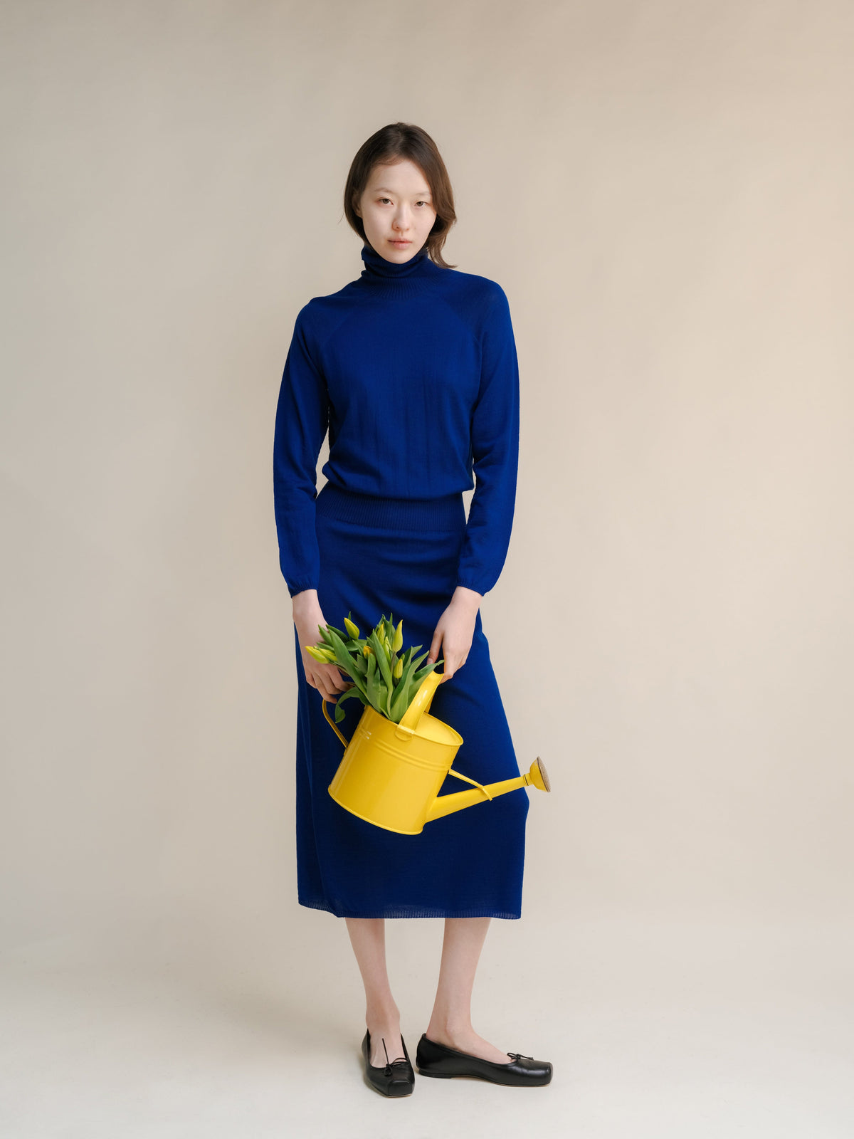 Model in a royal blue turtleneck dress holding a yellow watering can with green plants against a neutral backdrop