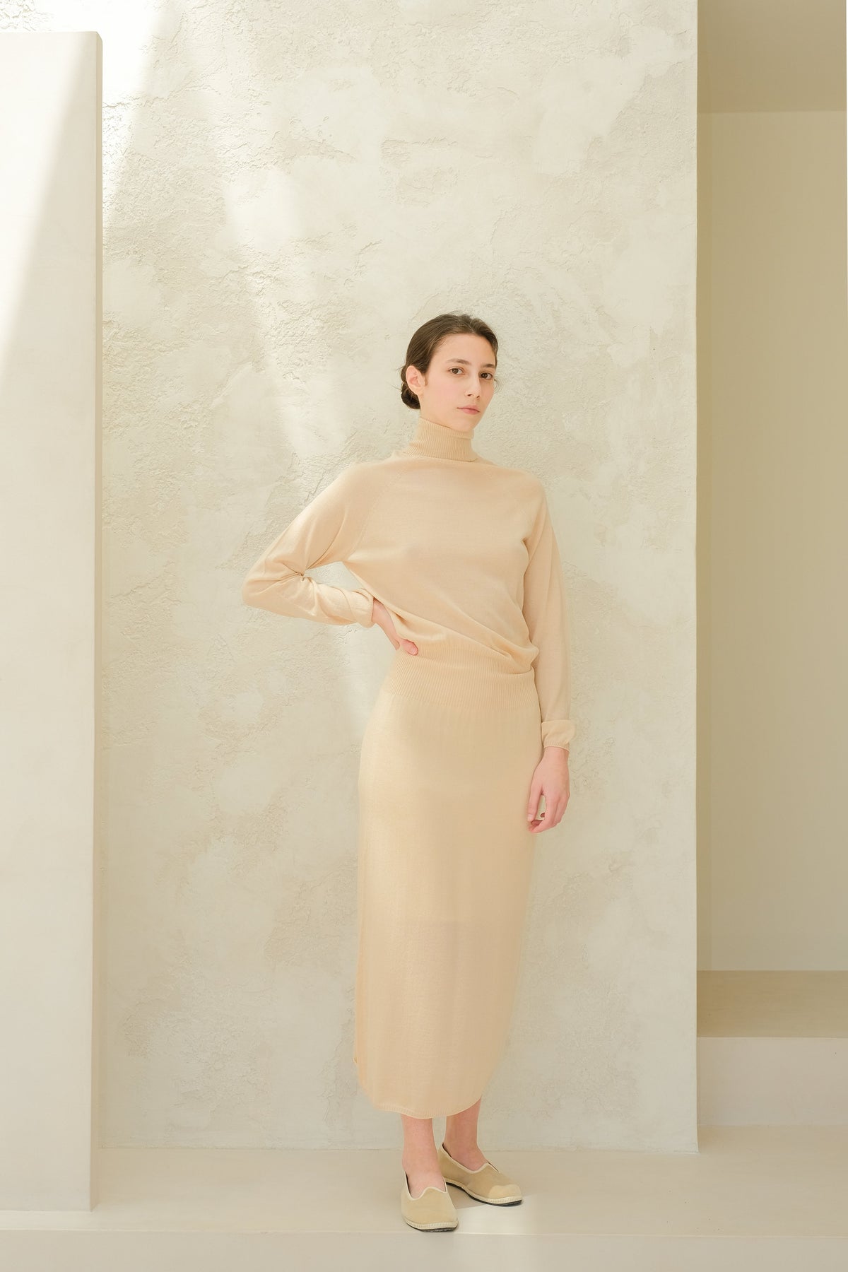 Full-length beige knit dress with a turtleneck sweater and matching skirt, model stands with hand on hip.