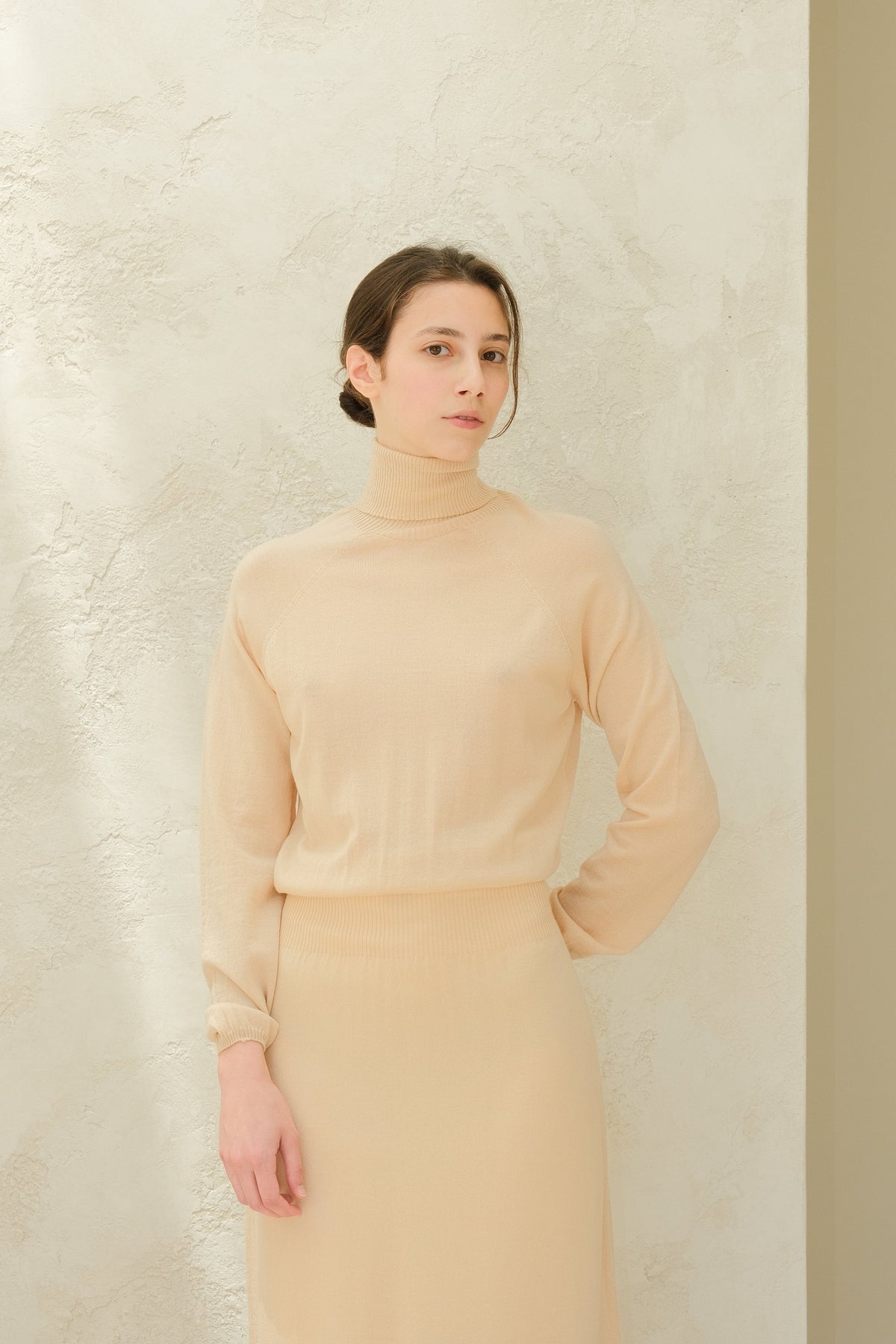 Beige knit turtleneck dress with a fitted waist and ankle-length skirt, model poses against textured wall.
