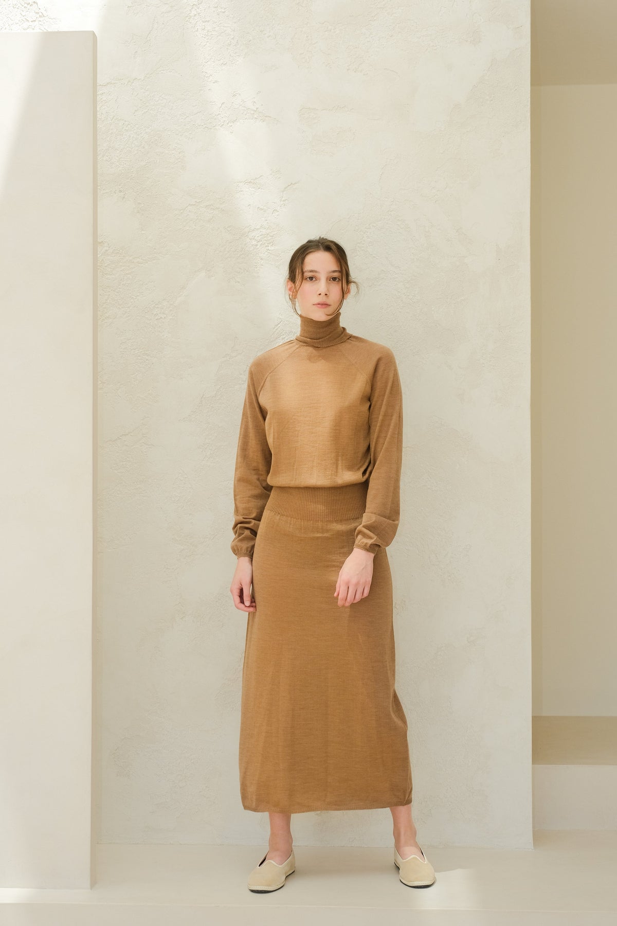 Full-body view of a model in camel brown 3D knit merino dress set with a turtleneck top and ribbed-waist skirt, beige slip-on shoes