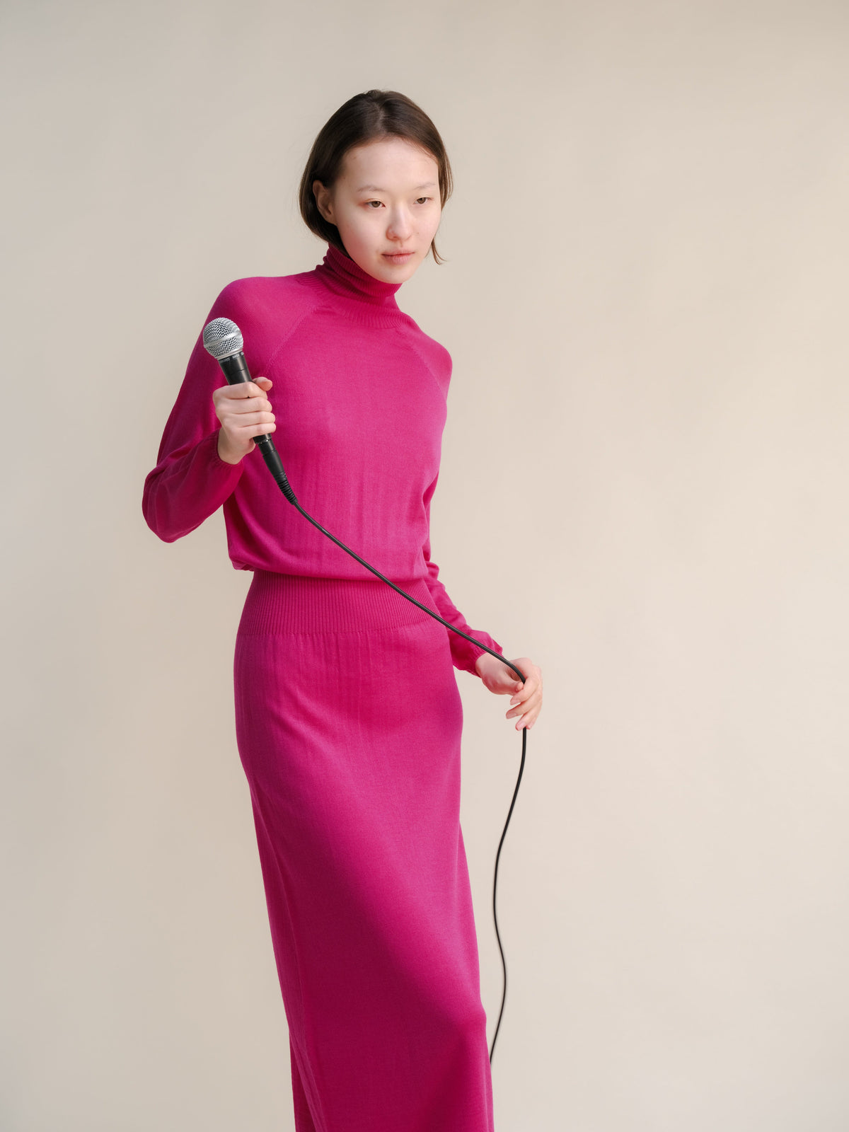 Model in magenta 3D knit merino wool dress holding a microphone against a pale studio backdrop