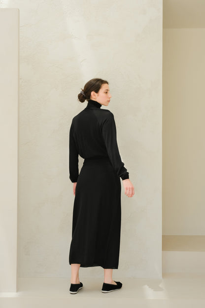 Back view of a model wearing a black 3D knit extra fine merino wool dress with a turtleneck and long sleeves, midi length