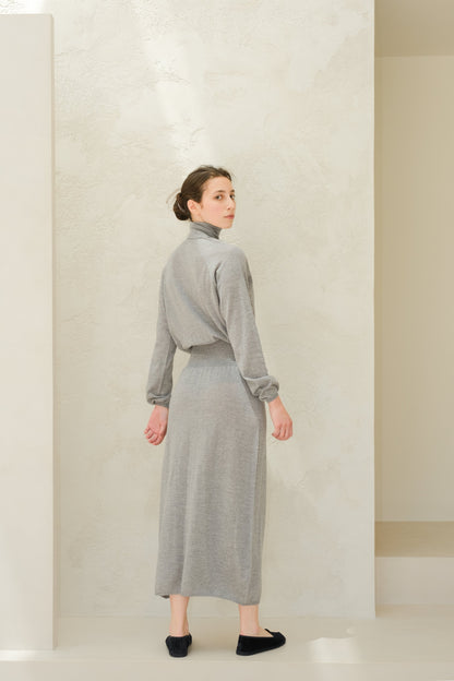 Model wearing a light grey 3D knit merino wool dress from behind with a cinched waist and long sleeves.