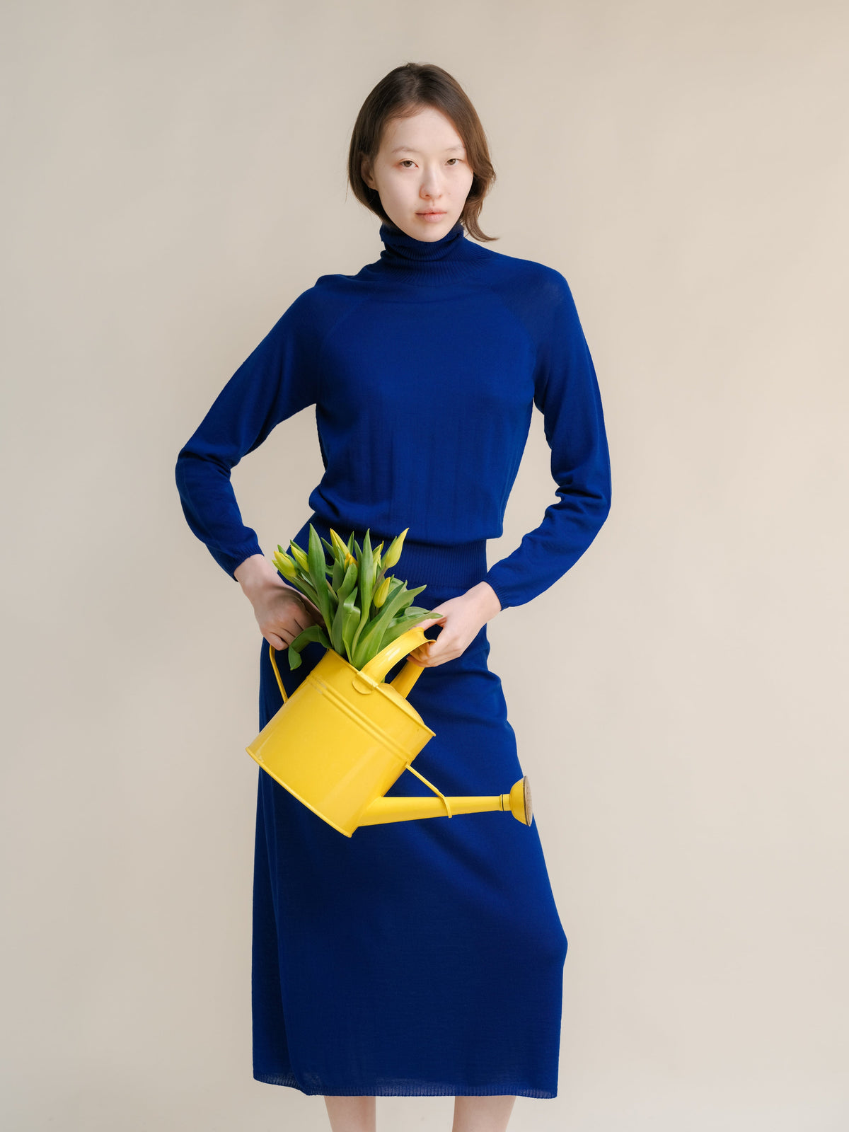 Blue 3D-Knit merino wool dress with turtleneck on a model holding a yellow watering can with tulips