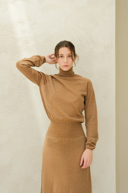 Front view of a model wearing a camel brown 3D knit merino wool dress with a turtleneck and ribbed waist against a pale textured wall