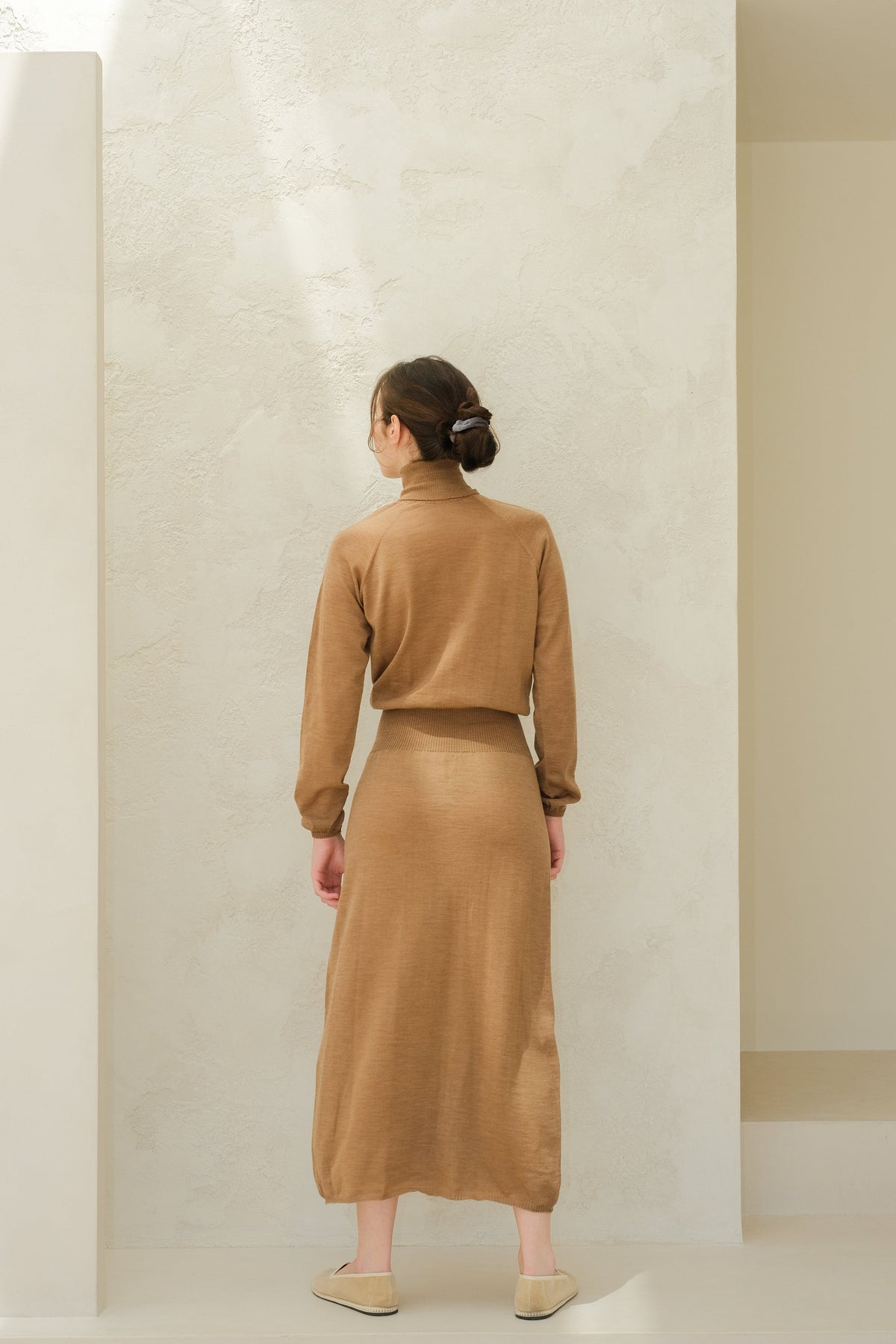 Back view of a model in camel brown knit dress with matching long skirt, hair in a bun.
