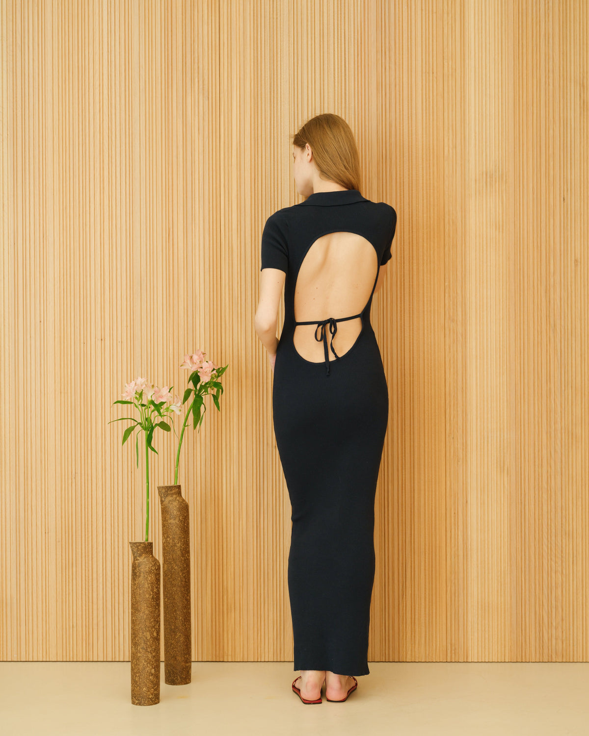 Back view of a mannequin porcognact a black open-back cotton dress with tie strings debout devant a wood panel wall.