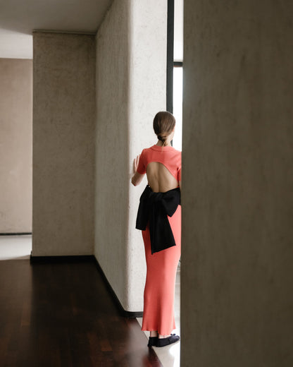 Back view of a model in a coral open-back dress with an oversized black bow tied at the lower back.