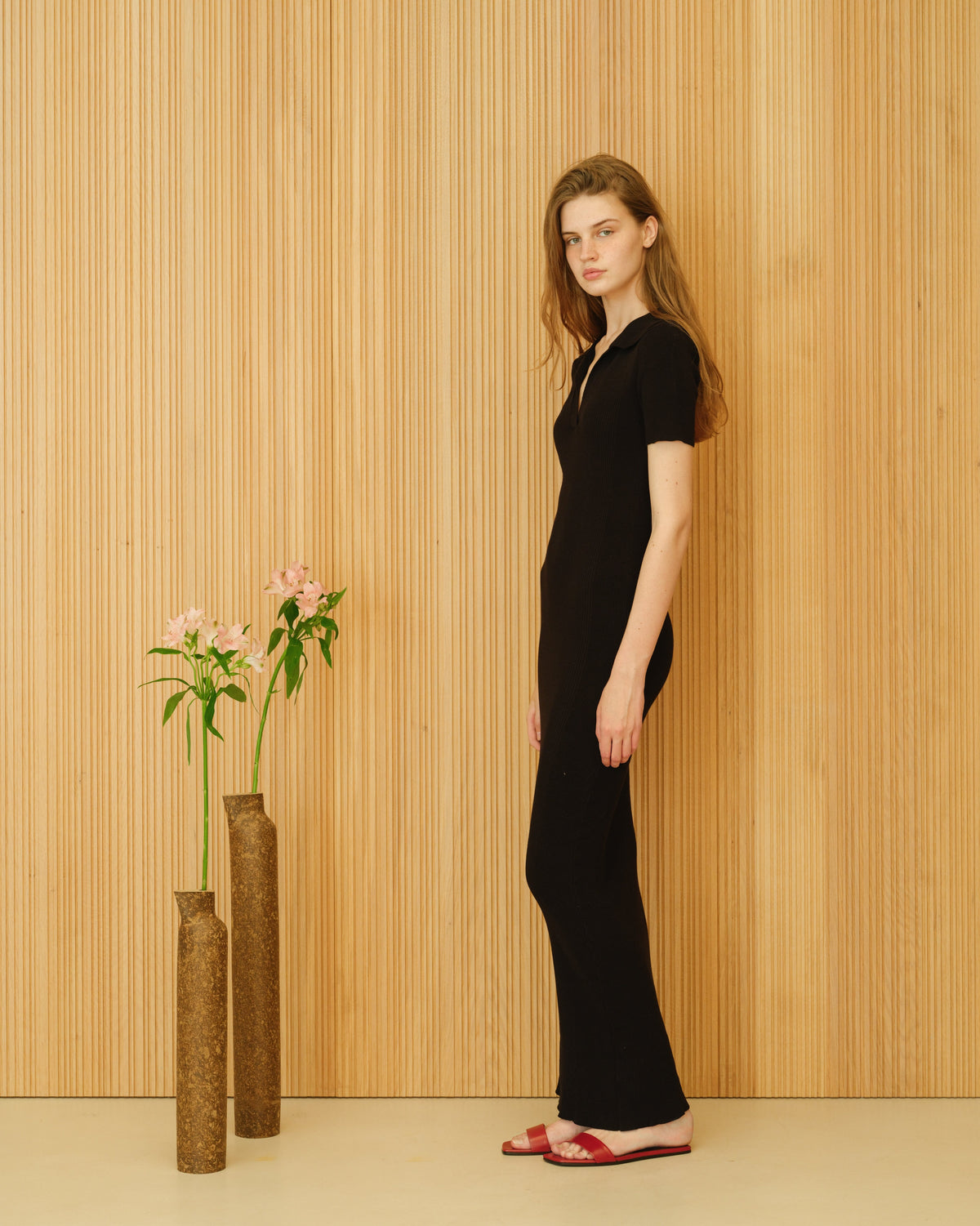 Mannequin en a black cotton dress scognacds in profile against a wood panel wall with brown vases and pink flowers.