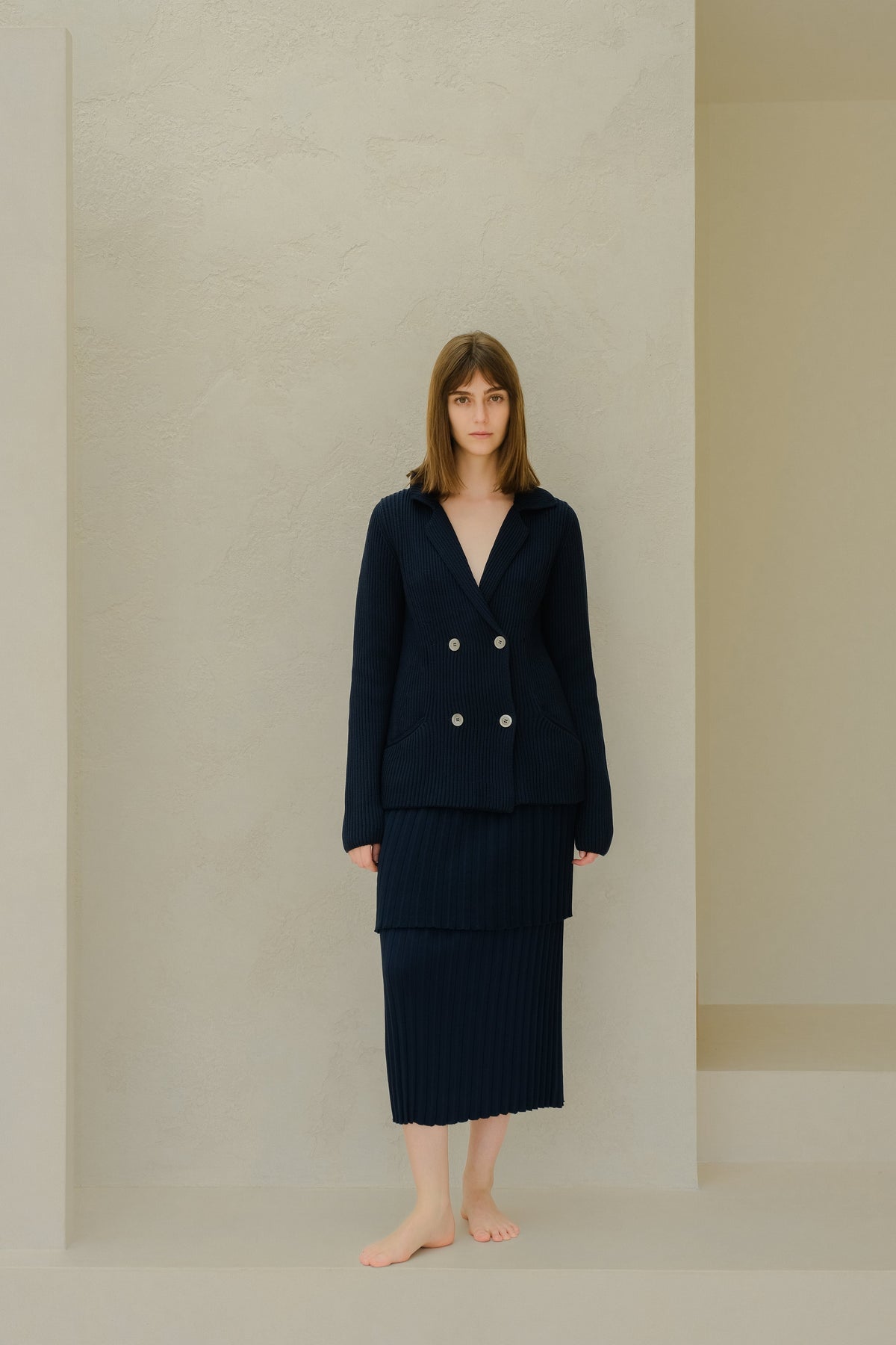 Navy knitted double breasted blazer with a layered pleated long skirt on a barefoot model against a beige textured wall