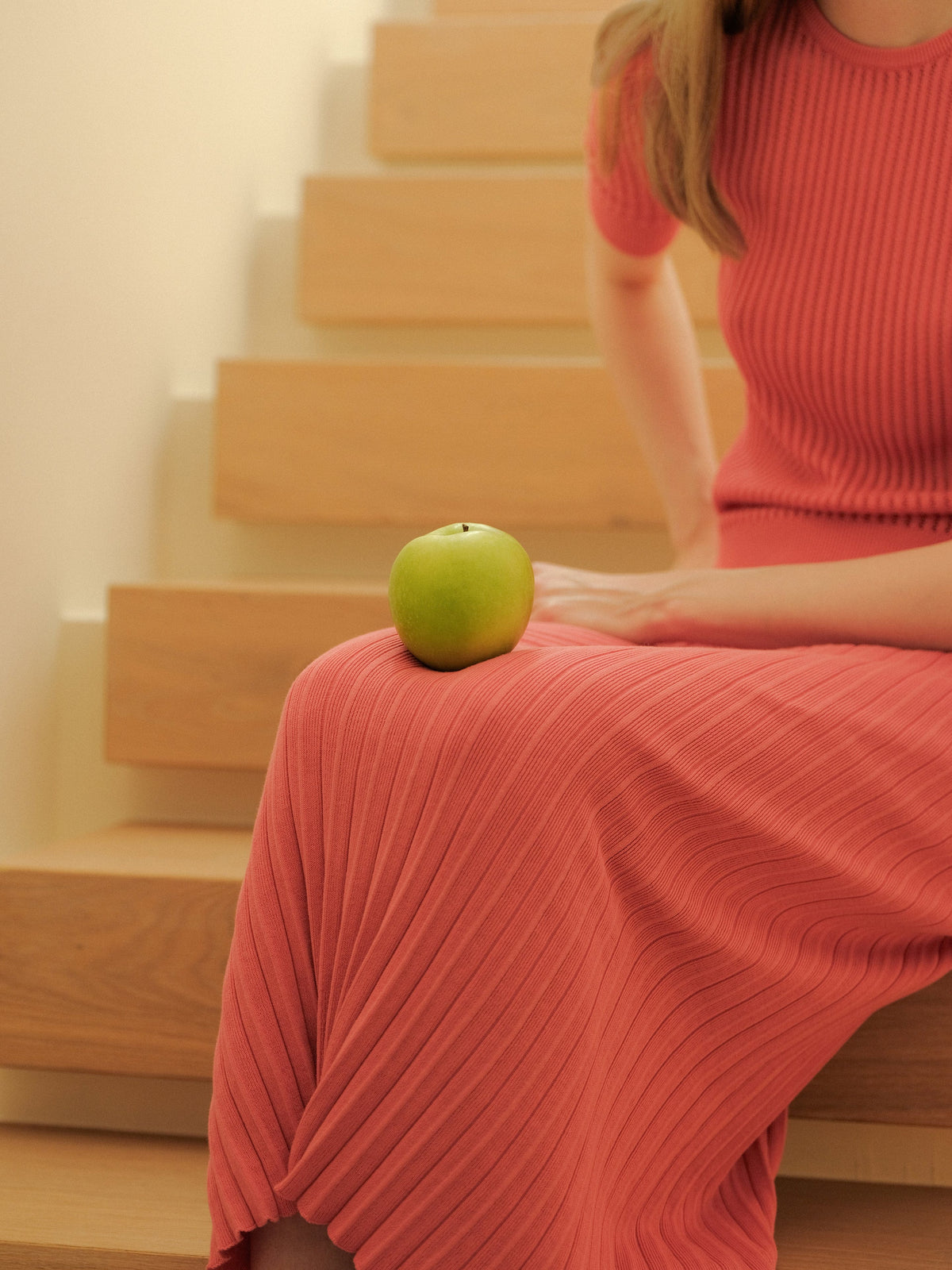 Green apple rests on a coral pink long pleated skirt worn by a model sitting on wooden stairs.