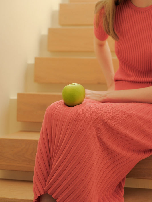 Green apple rests on a coral pink long pleated skirt worn by a model sitting on wooden stairs.