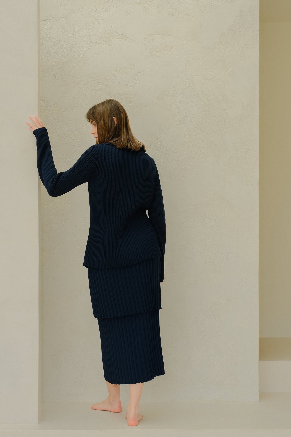 Back view of a model wearing a navy knit plissé skirt set against a beige wall.