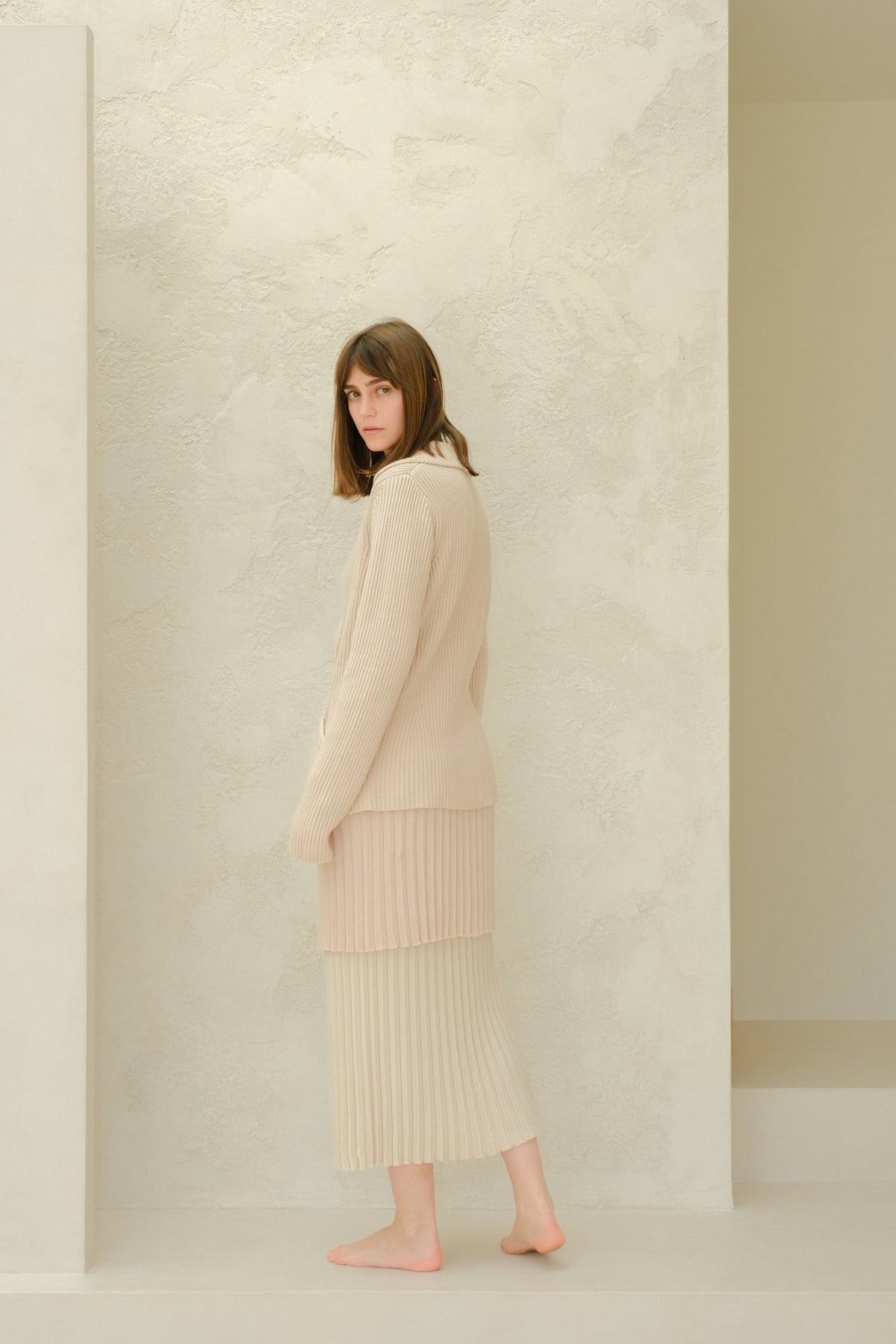 Side view of a model in a beige Fine Cotton Plissé knit ensemble with a long pleated skirt