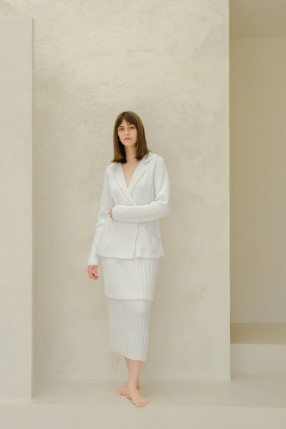 Side view of the model in a pale gray knit blazer and matching pleated long skirt against a textured light wall.