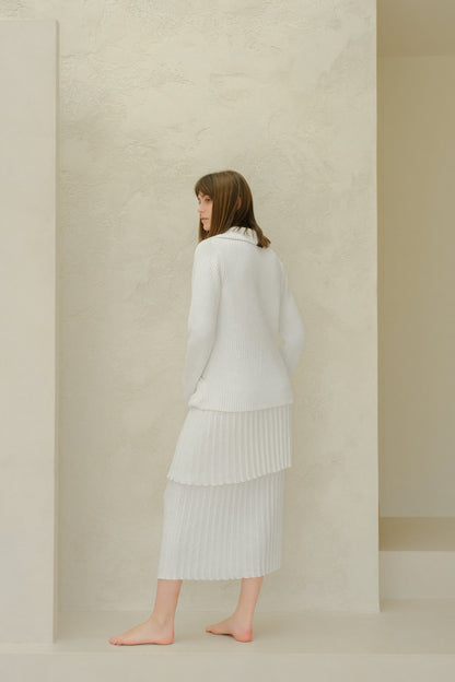 Side view of a model wearing a white fine cotton plissé knit skirt with a matching sweater against a textured beige wall.