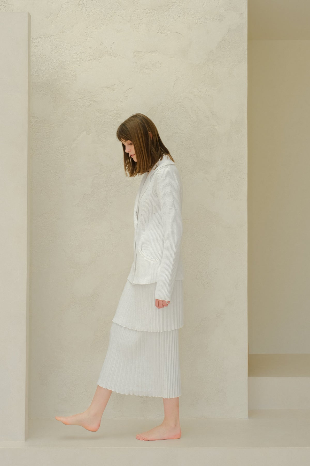 Side profile of a model in a white knitted blazer with a layered pleated long skirt from Fine Cotton Plissé