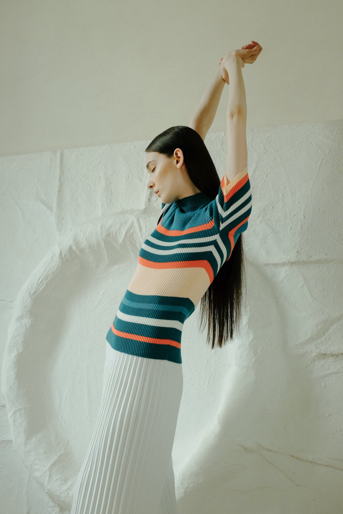 Profile of a model in a multicolor striped knit top and a white pleated long skirt, arms raised overhead.