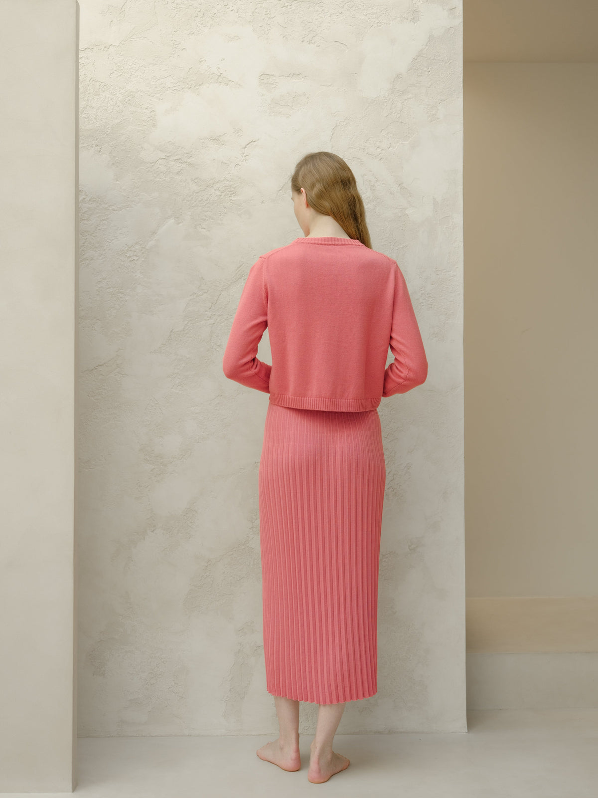 Back view of coral knit cropped cardigan with a matching long pleated skirt worn by a barefoot model.