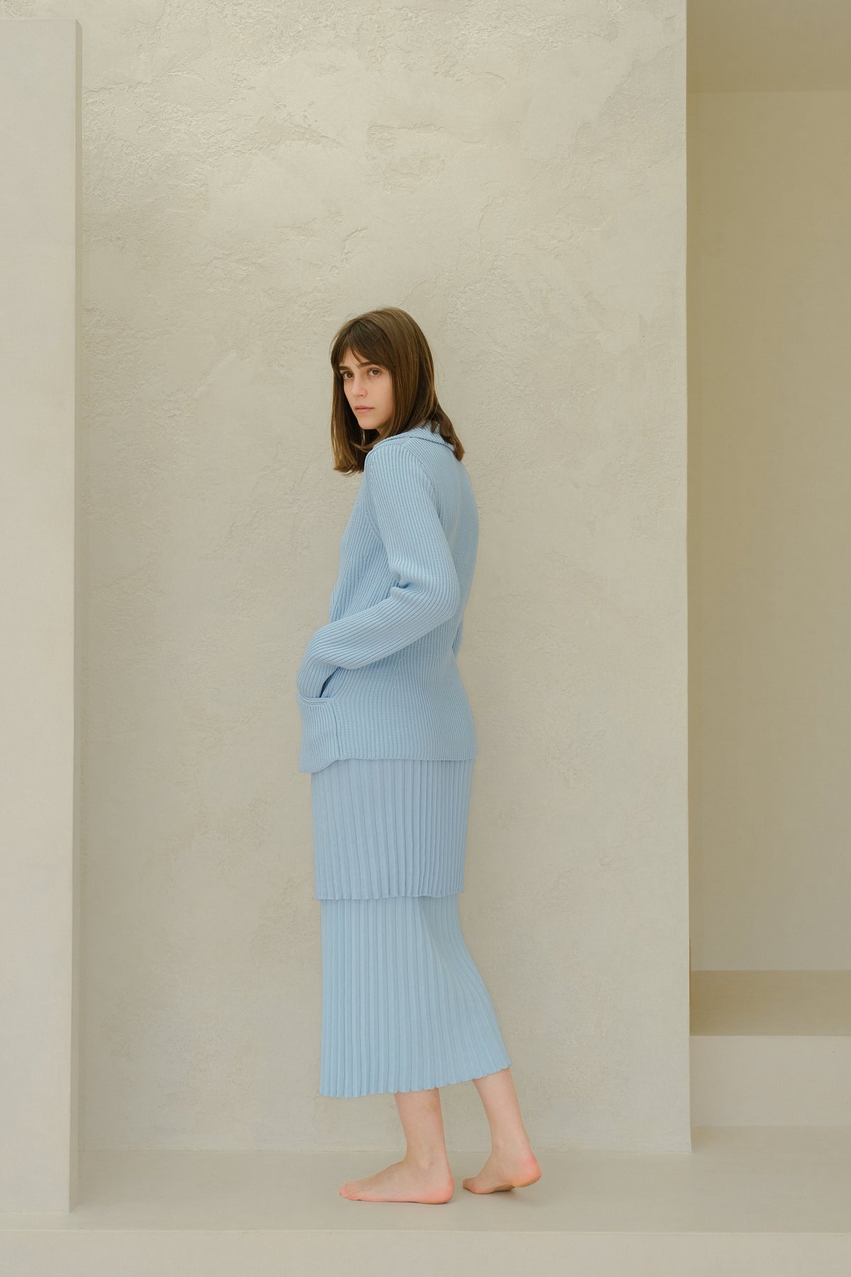 Side view of a model wearing a light blue Fine Cotton Plissé Long Skirt with a matching knit sweater in a neutral studio setting.