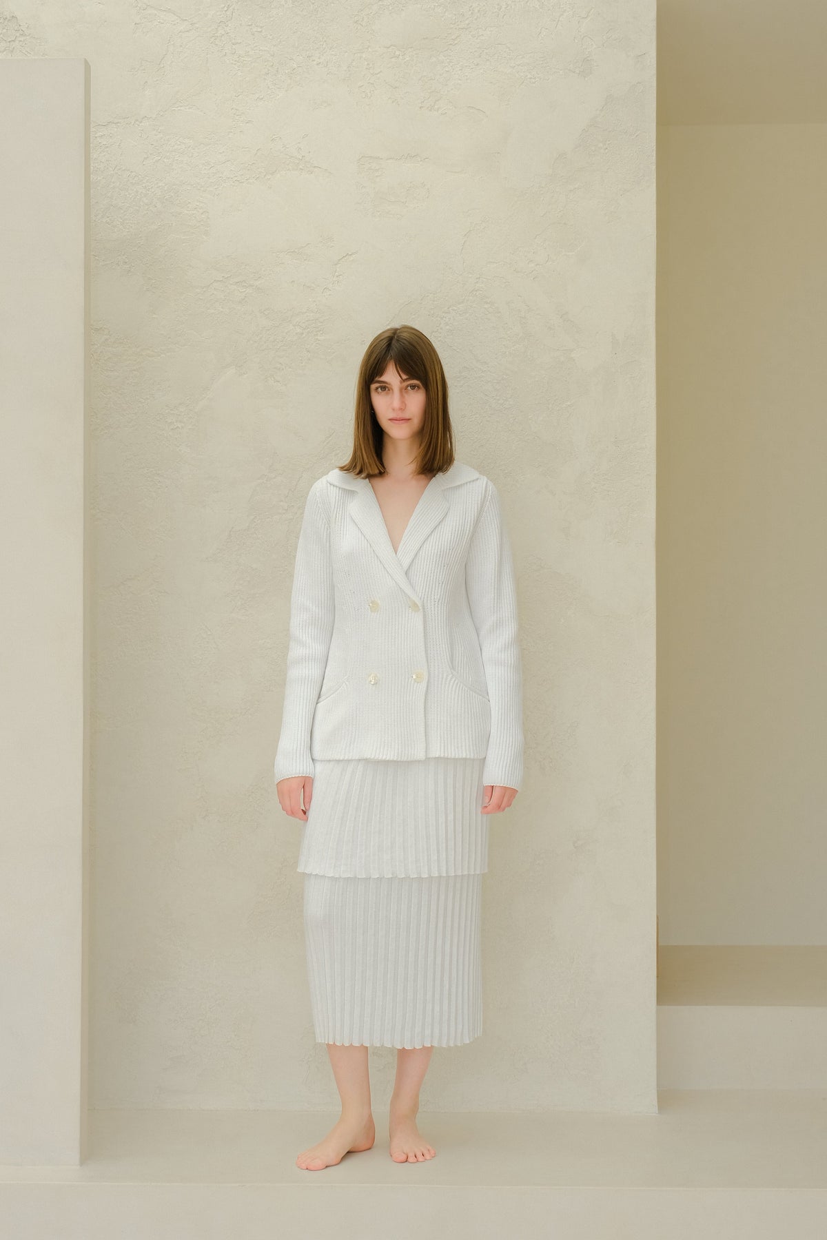 Model wearing a light knit double-breasted blazer and pale pleated long skirt against a neutral studio wall.