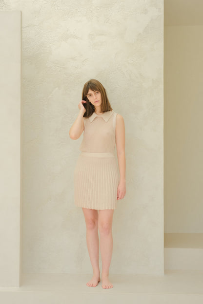 Modella con a beige fine cotton plissé minigonna e top in maglia senza maniche davanti a una parete chiara testurizzata.