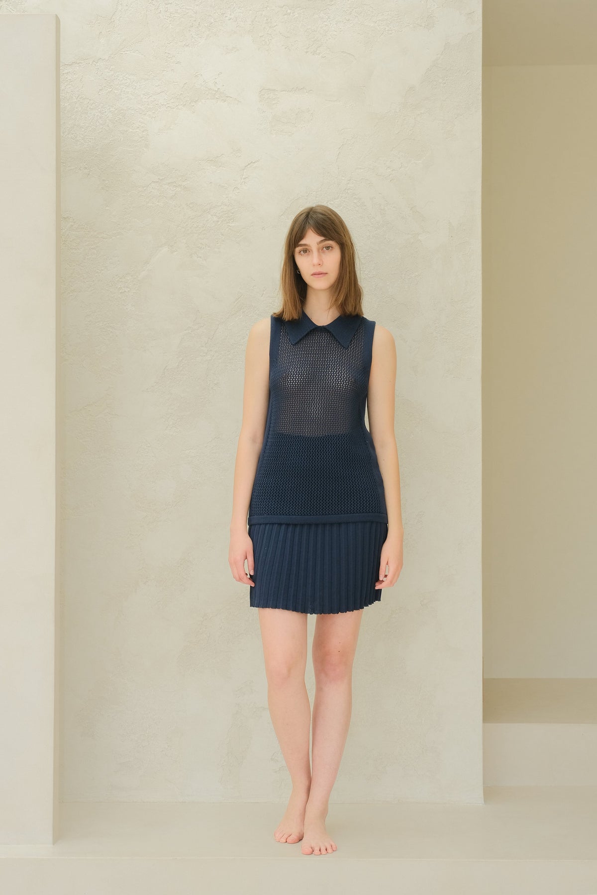 Vista frontale a figura intera of a modella con a blu navy sleeveless top in maglia traforata con a colletto a punta e a pleated minigonna.