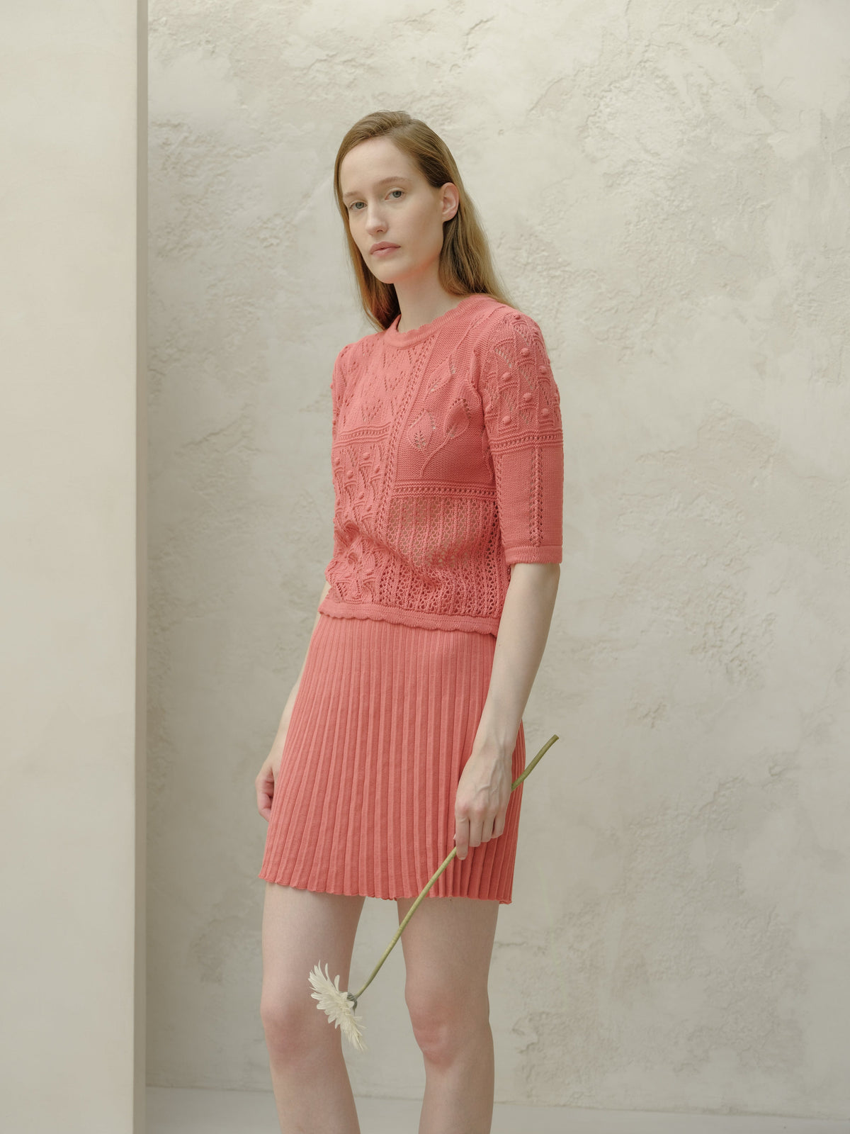 Modella con rosa minigonna plissé in cotone fine con a top in maglia coordinato stes davanti a una parete chiara testurizzata, con un fiore.