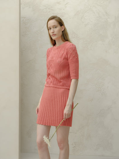 Modella con rosa minigonna plissé in cotone fine con a top in maglia coordinato stes davanti a una parete chiara testurizzata, con un fiore.
