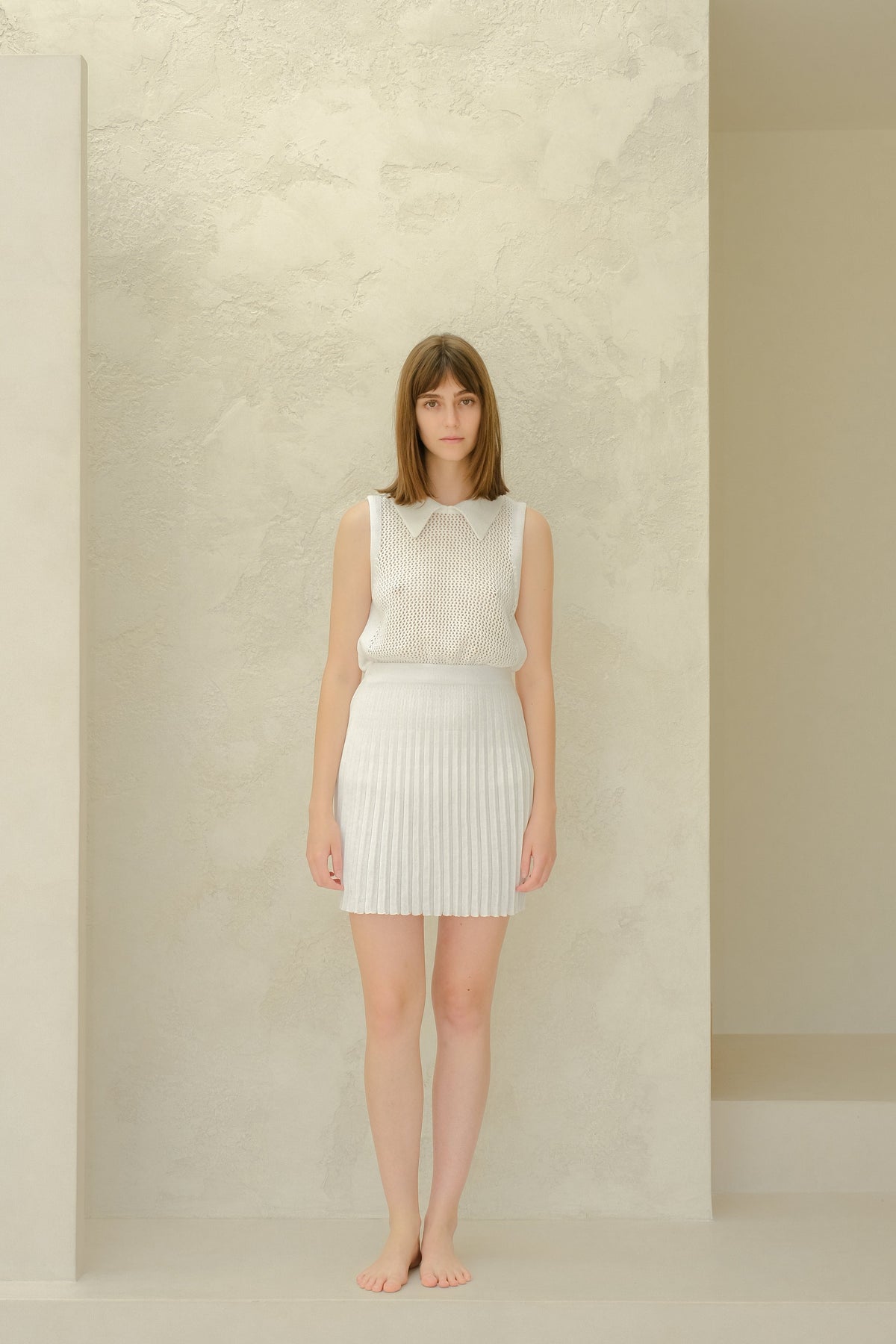 Figura intera of a modella con a bianco mesh sleeveless top con a collarosso neckline e a bianco plissé minigonna, barefoot.