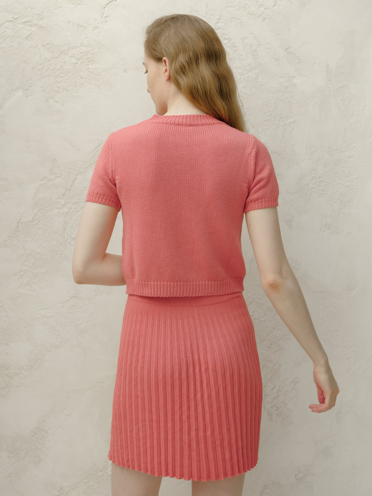 Back view of a modella con a coral rosa knitted top e matching pleated minigonna