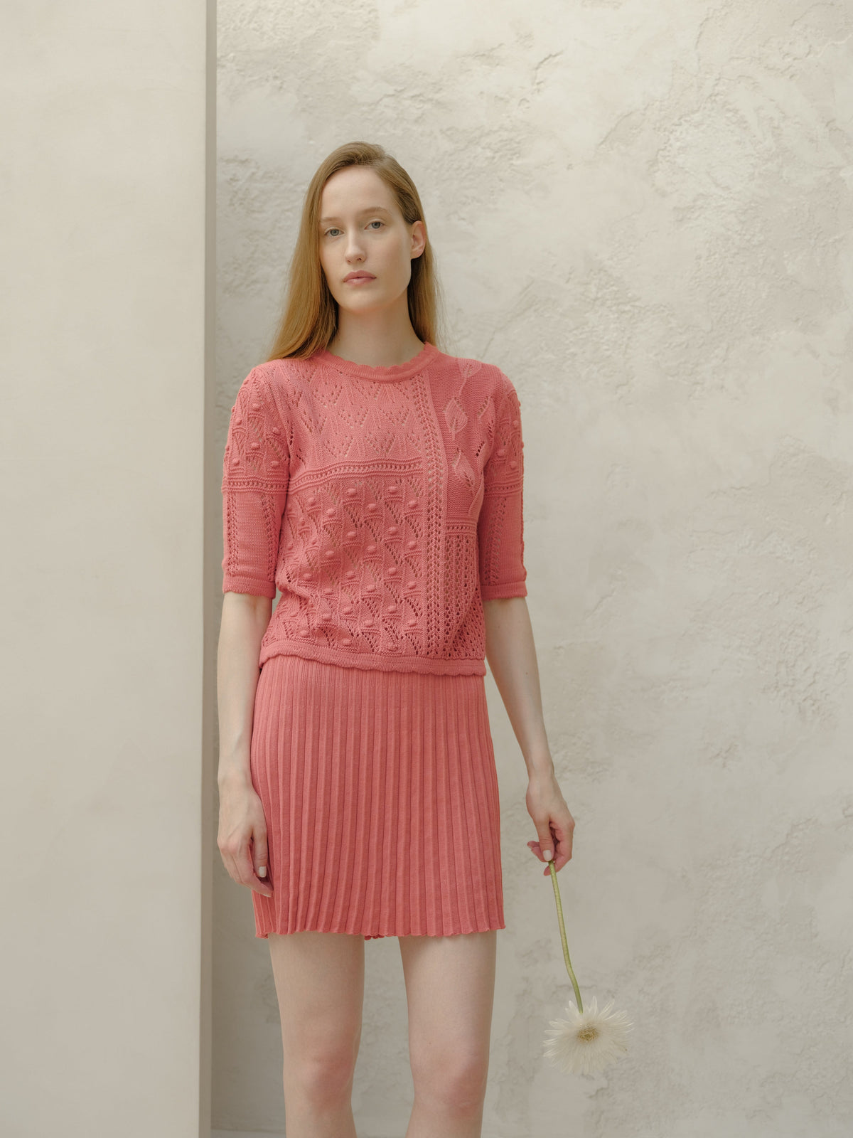 Vista frontale of a model in a rosa minigonna plissé in cotone fine e top in maglia coordinato davanti a una parete chiara testurizzata.