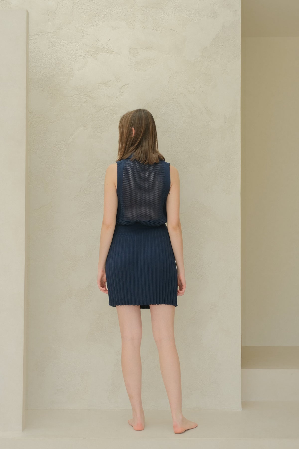 Back view of a model in a blu navy sleeveless top in maglia traforata con a colletto a punta e pleated minigonna.