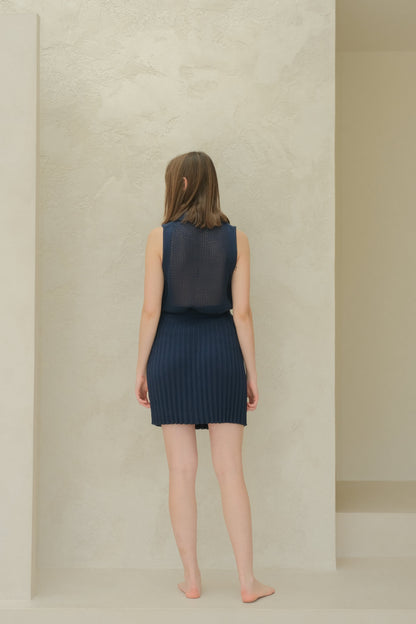 Back view of a model in a blu navy sleeveless top in maglia traforata con a colletto a punta e pleated minigonna.